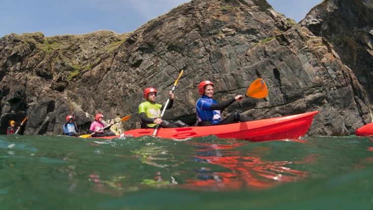 Several people in tandem kayaks are paddling in the water at Mullion Cove, Cornwall.